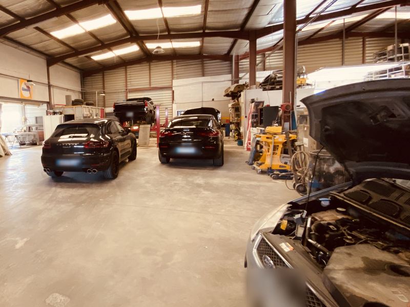 Intérieur de l'atelier STELO MAP : voitures garées sous la charpente métallique, capot ouvert au premier plan, plaques floutées